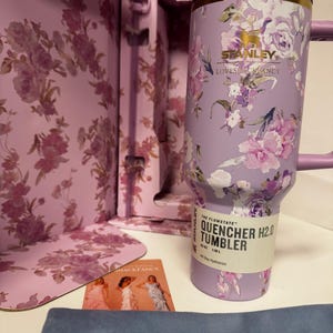May include: A lavender Stanley Quencher H2.0 tumbler with a floral pattern, gold rim, and purple lid. The label reads The Flowstate Quencher H2.0 Tumbler 40 oz 1.18 L. It is presented in a matching floral box with a small card featuring three figures.