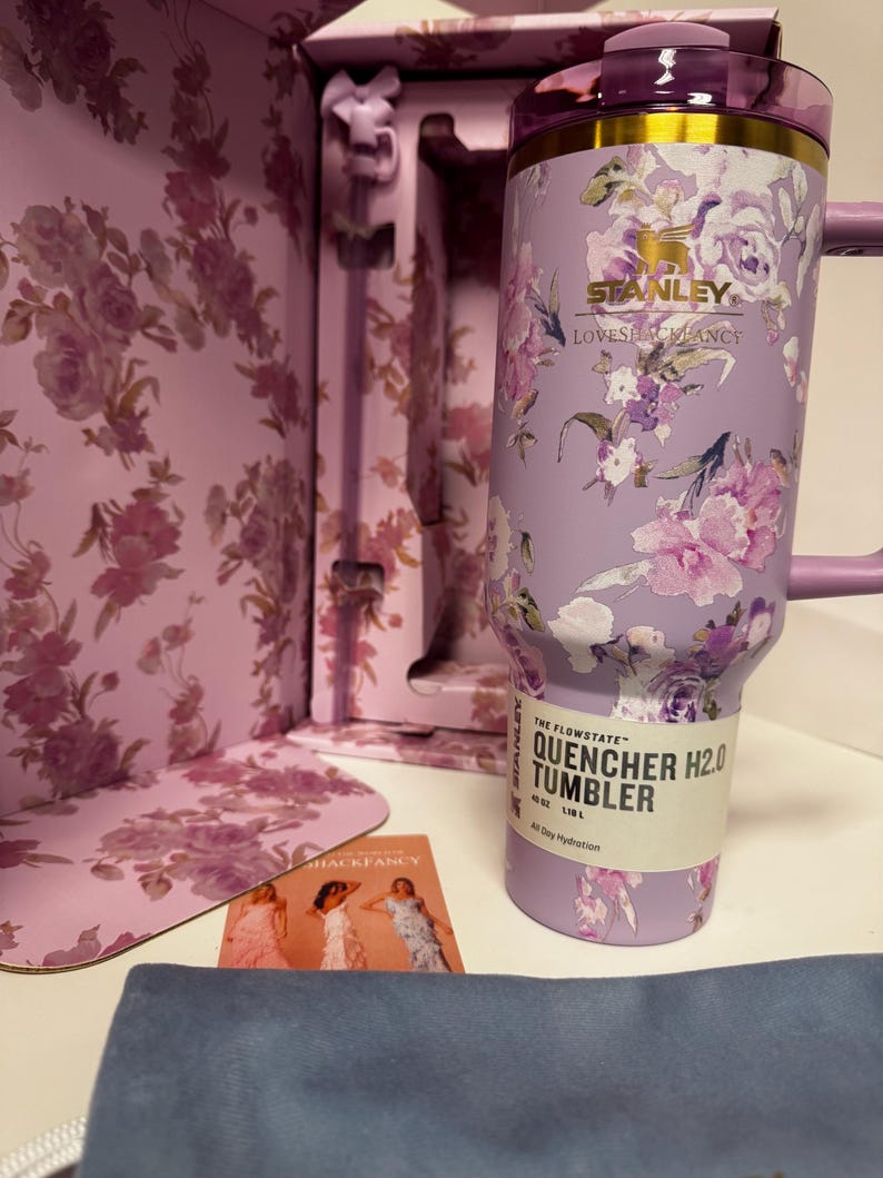 May include: A lavender Stanley Quencher H2.0 tumbler with a floral pattern, gold rim, and purple lid. The label reads The Flowstate Quencher H2.0 Tumbler 40 oz 1.18 L. It is presented in a matching floral box with a small card featuring three figures.