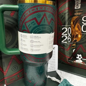 May include: A dark green stainless steel tumbler with a gold rim and a handle, featuring a red and green geometric pattern. A white paper band wraps around the center, displaying product icons and text. The tumbler is set against a matching box.