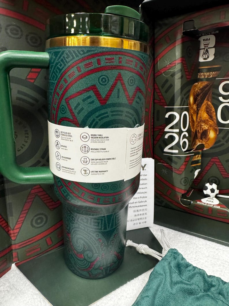 May include: A dark green stainless steel tumbler with a gold rim and a handle, featuring a red and green geometric pattern. A white paper band wraps around the center, displaying product icons and text. The tumbler is set against a matching box.