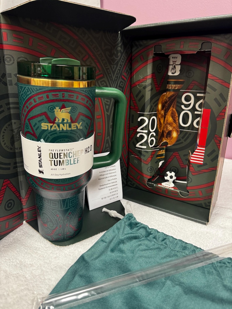 May include: A dark green Stanley Quencher H2.0 40 oz tumbler with a gold rim and green lid, featuring a red and green geometric pattern. The tumbler is inside a box with FIFA World Cup 2026 branding, accompanied by a green fabric pouch and a clear straw.