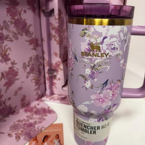 May include: A lavender Stanley Quencher H2.0 tumbler featuring a floral pattern, a gold-tone rim, and a purple lid. The tumbler is displayed in front of its matching floral-patterned box and includes a small card showing three dresses.