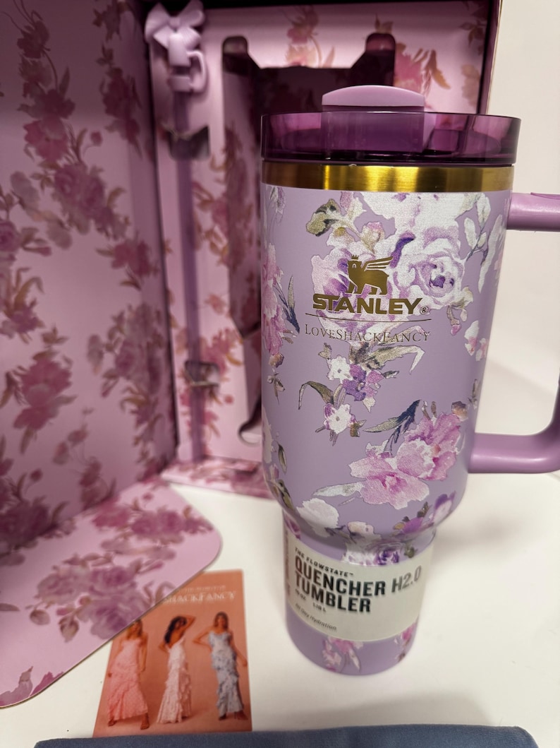 May include: A lavender Stanley Quencher H2.0 tumbler featuring a floral pattern, a gold-tone rim, and a purple lid. The tumbler is displayed in front of its matching floral-patterned box and includes a small card showing three dresses.