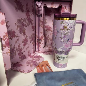 May include: A lavender Stanley Quencher H2.0 FlowState tumbler with a floral pattern, a matching straw, and a purple lid. The tumbler is inside a floral-patterned box with a blue velvet drawstring pouch and a small card featuring three figures.