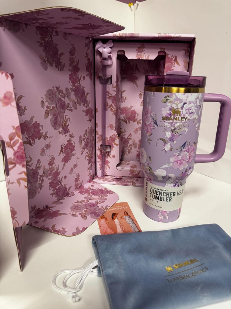 May include: A lavender Stanley Quencher H2.0 FlowState tumbler with a floral pattern, a matching straw, and a purple lid. The tumbler is inside a floral-patterned box with a blue velvet drawstring pouch and a small card featuring three figures.