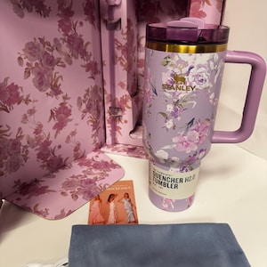 May include: A lavender Stanley Quencher H2.0 tumbler with a floral pattern, a gold-tone rim, and a matching handle. The tumbler is inside a floral-patterned box with a bow-shaped straw topper, a small informational card, and a blue drawstring pouch.