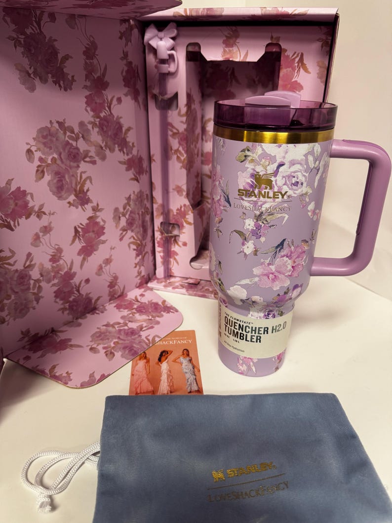 May include: A lavender Stanley Quencher H2.0 tumbler with a floral pattern, a gold-tone rim, and a matching handle. The tumbler is inside a floral-patterned box with a bow-shaped straw topper, a small informational card, and a blue drawstring pouch.