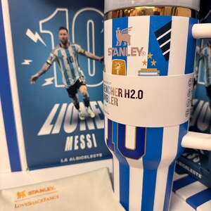 Może przedstawiać: Niebiesko-biały kubek Stanley Quencher H2.0 w paski ze złotą krawędzią, białym uchwytem i papierową opaską z napisem Stanley Quencher H2.0 Tumbler. Kubek ozdobiony jest grafiką z numerem 10 oraz ikonami związanymi z piłką nożną.