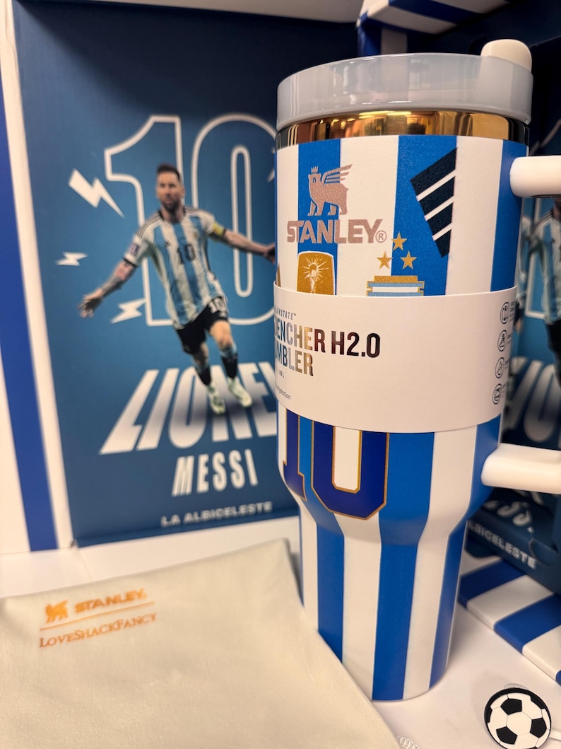 Może przedstawiać: Niebiesko-biały kubek Stanley Quencher H2.0 w paski ze złotą krawędzią, białym uchwytem i papierową opaską z napisem Stanley Quencher H2.0 Tumbler. Kubek ozdobiony jest grafiką z numerem 10 oraz ikonami związanymi z piłką nożną.