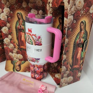 May include: A white Stanley Quencher H2.0 tumbler featuring a pink handle and lid, decorated with a floral and Our Lady of Guadalupe print. The tumbler is displayed with its matching box, a pink fabric pouch, and a small pink charm with the text I do.