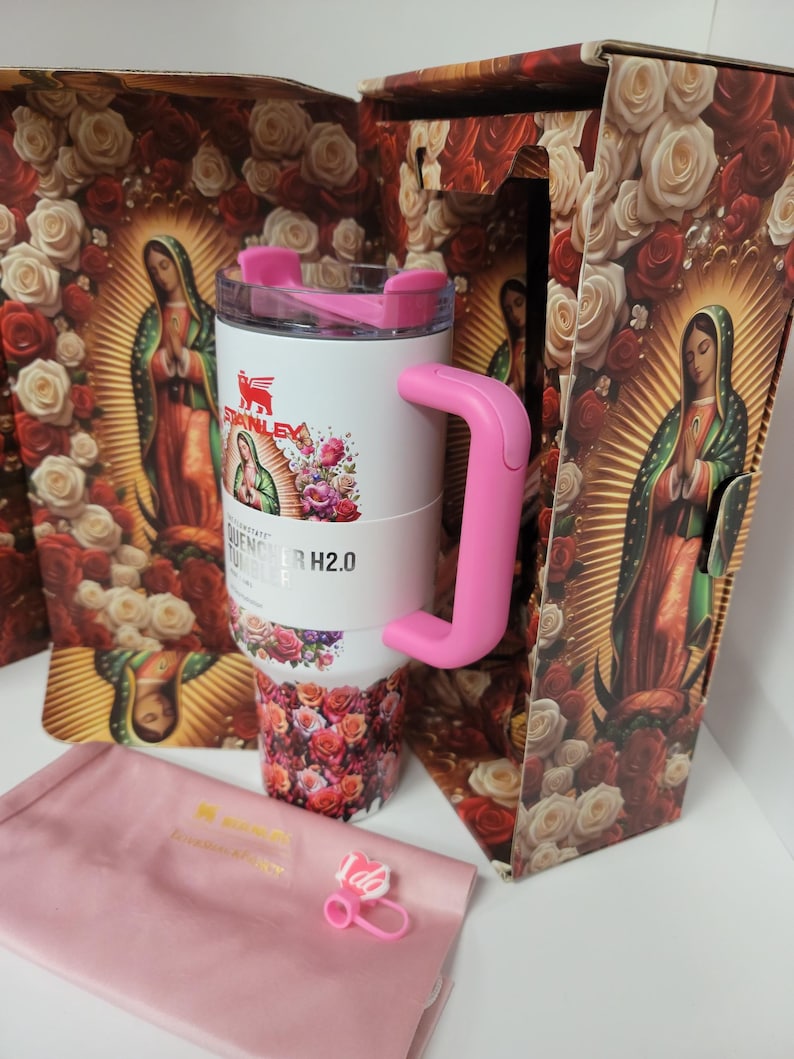 May include: A white Stanley Quencher H2.0 tumbler featuring a pink handle and lid, decorated with a floral and Our Lady of Guadalupe print. The tumbler is displayed with its matching box, a pink fabric pouch, and a small pink charm with the text I do.