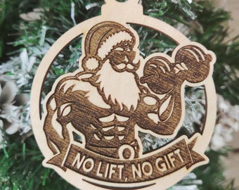 May include: A wooden Christmas ornament shaped like a circle with a cartoon image of Santa Claus lifting a dumbbell. The ornament has the text "No Lift, No Gift" on a banner.