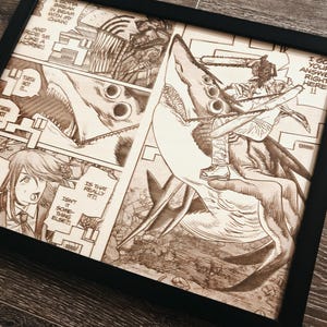 May include: Framed artwork featuring a detailed manga panel with a chainsaw-wielding character riding a shark-like creature. The artwork is in shades of brown and black, with text bubbles and action lines. The frame is black.