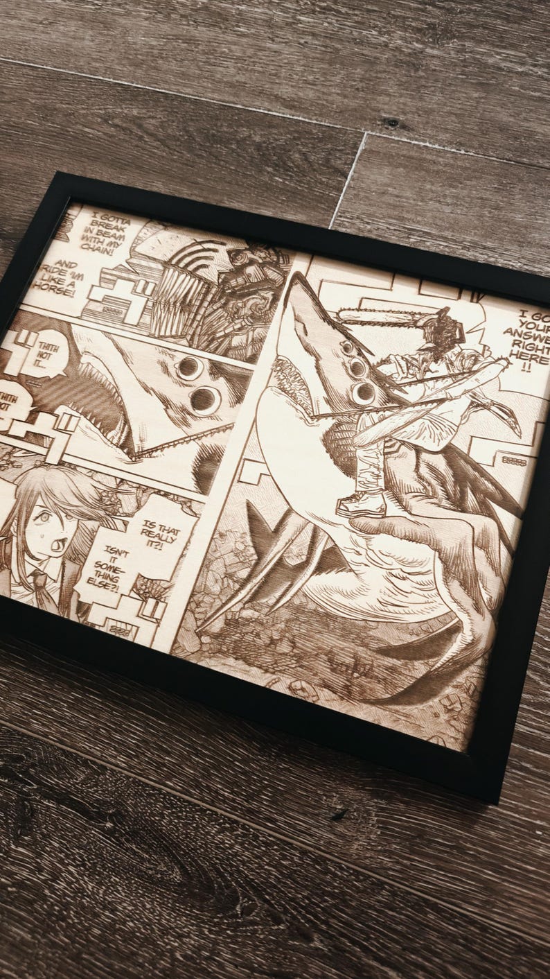 May include: Framed artwork featuring a detailed manga panel with a chainsaw-wielding character riding a shark-like creature. The artwork is in shades of brown and black, with text bubbles and action lines. The frame is black.