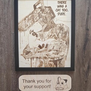 May include: Wooden framed artwork with a detailed laser-etched illustration of a chainsaw-wielding character and a cat. The text "THERE WAS A CAT TOO, DUDE." is visible. A small wooden sign below reads "Thank you for your support!"