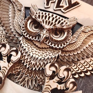 May include: A wooden owl with intricate laser-cut details, its wings spread wide. The owl's eyes are large and round, and its feathers are detailed with a variety of patterns. The word "FLY" is carved above the owl.