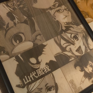 May include: A framed wood panel featuring engraved anime-style illustrations. The artwork includes various characters with expressive faces and Japanese text. The frame is black, and the wood has a natural, light brown color.