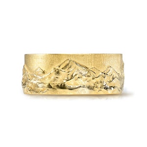 May include: A gold band ring with a mountain range design. The ring is wide and has a textured surface.