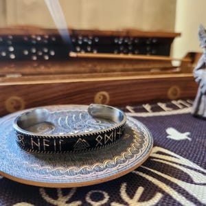 Puede incluir: Una pulsera de metal negro con runas y símbolos plateados. La pulsera está sobre un posavasos de madera con un diseño de olas.