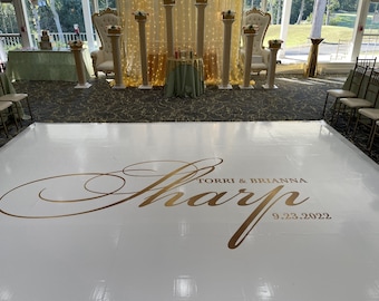 May include: A white dance floor with gold lettering that reads "Sharp" and the names "Torri & Brianna" with the date "9.23.2022". The background includes wedding decorations, chairs, and a table.