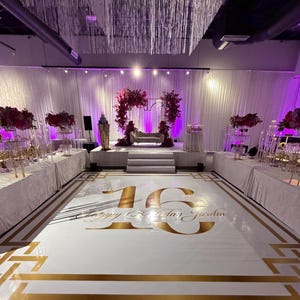 May include: An event space decorated for a celebration. The white floor features gold accents and the text "16 Happy Birthday Jardin." A stage is set with a couch, floral arrangements, and a backdrop of white curtains. Overhead, crystal strands hang from the ceiling.