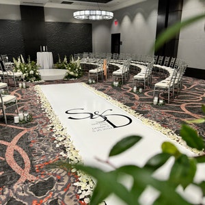 May include: A white aisle runner with white flower petals lining the sides. The runner has the couple's initials and names, "Sherri and Derrick", and the date "5.28.2022" printed in black. The aisle runner is in a wedding ceremony space with white chairs arranged in a semi-circle.