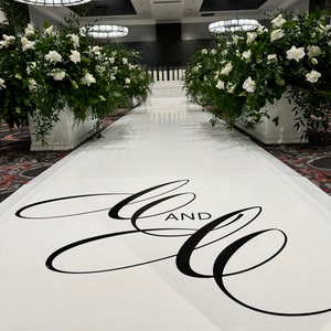 May include: A white aisle runner with the words "and" and two stylized cursive "C"s in black. The aisle runner is in a large room with white walls and white floral arrangements on either side.