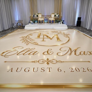 May include: A large, rectangular, cream-colored dance floor featuring gold-colored lettering and a circular monogram. The text reads "Ella & Max" and "AUGUST 6, 2026" in a metallic finish. A gold-colored floral wreath surrounds the "EM" monogram.