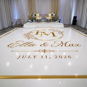 May include: A white glossy dance floor featuring a gold monogram with the letters EM inside a circular wreath, the names Ella & Max in gold script, and the date JULY 11, 2026, all framed by a thin gold border on the rectangular surface.