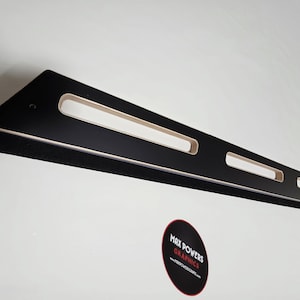 May include: A black wooden shelf with three rectangular cutouts and a black felt strip along the bottom edge. The shelf is mounted on a white wall. A circular sticker with the text "MAX POWERS GRAPHICS" is visible.