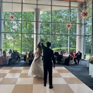 May include: A bride and groom dance on a checkered dance floor in a large room with floor-to-ceiling windows. Guests are seated at tables in the background.