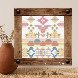 May include: A framed cross-stitch with a Southwestern design. The artwork features geometric patterns in shades of pink, blue, yellow, and orange. The frame is made of rustic wood and has dark metal accents. The text "Cotton Valley Stitches" is at the bottom.