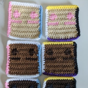 May include: Six handmade crochet coasters in a square shape, resembling slices of cake. Each coaster features a different color scheme, including pink, blue, brown, and yellow, with a white border and a small pink heart design.