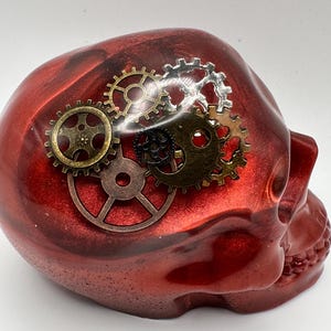 May include: A red skull sculpture with a metallic, glittery finish. The skull is adorned with several bronze and silver-colored gears, creating a steampunk aesthetic. The sculpture is a unique decorative item.