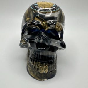 May include: A decorative skull sculpture with a gold-colored jaw and accents. The skull has a clear, transparent body with black and gold swirls. The top of the skull features a gold emblem with the text "WELL AND BRING THE MEETING".