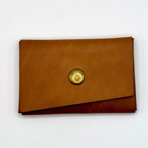 May include: A small, rectangular, brown leather wallet with a brass-coloured snap closure. The wallet is closed, with a flap folded over the top. The leather has a smooth texture and a warm, natural colour.