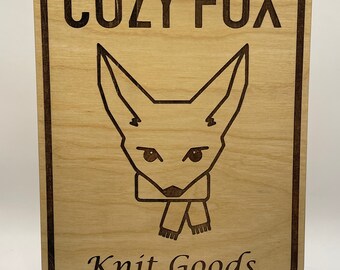 May include: Wooden sign with the text "COZY FOX" and "Knit Goods" engraved in a dark brown colour. The sign features a fox head illustration with a scarf. The sign is displayed on a wooden stand.