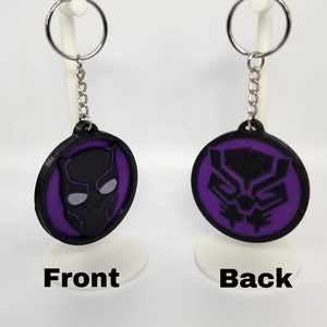Może przedstawiać: Brelok do kluczy Black Panther z fioletowym tłem i czarną sylwetką maski Black Panther. Tył breloka ma czarną sylwetkę maski Black Panther na fioletowym tle.