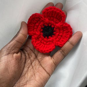 May include: A handmade, red crochet poppy flower with a black center. The flower has five petals and is made of a textured yarn. This floral accessory is a symbol of remembrance and can be used as a brooch or decorative item.