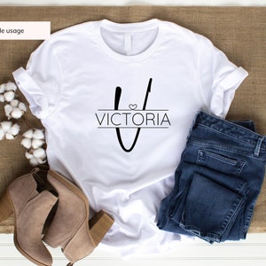 Peut inclure: T-shirt blanc avec un motif noir en forme de lettre "V" surmont&eacute;e d'un c&oelig;ur et du mot "VICTORIA" en dessous. Le t-shirt est pr&eacute;sent&eacute; sur un fond en toile de jute brune avec une paire de bottines marron &agrave; cheville, un jean bleu et des boules de coton.