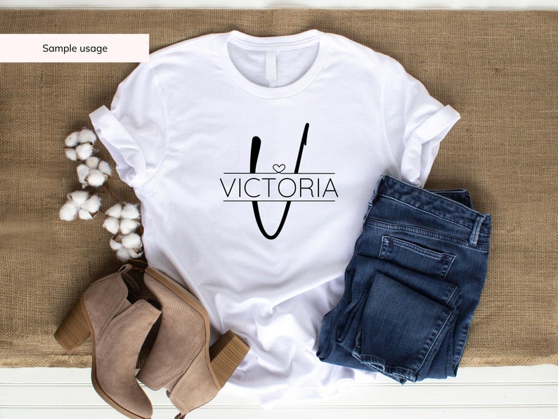 Peut inclure: T-shirt blanc avec un motif noir en forme de lettre "V" surmont&eacute;e d'un c&oelig;ur et du mot "VICTORIA" en dessous. Le t-shirt est pr&eacute;sent&eacute; sur un fond en toile de jute brune avec une paire de bottines marron &agrave; cheville, un jean bleu et des boules de coton.