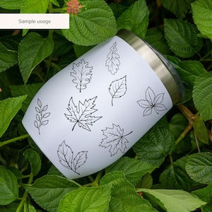 May include: White stainless steel tumbler with a black outline of various leaves. The tumbler is sitting on a bed of green leaves.