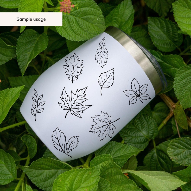 May include: White stainless steel tumbler with a black outline of various leaves. The tumbler is sitting on a bed of green leaves.