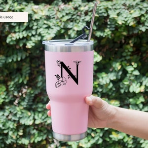 May include: A pink stainless steel tumbler with a black floral design and the letter "N" on the side. The tumbler has a lid and a straw.