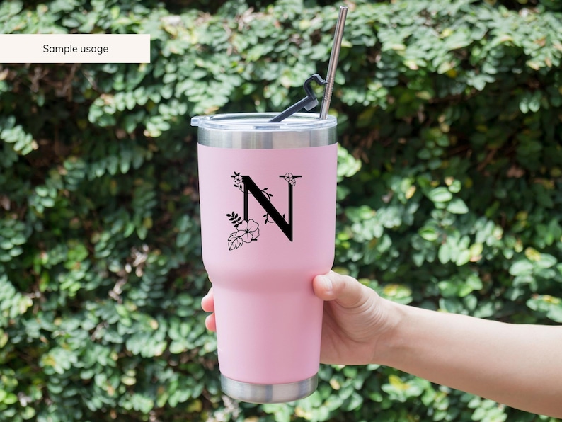 May include: A pink stainless steel tumbler with a black floral design and the letter "N" on the side. The tumbler has a lid and a straw.