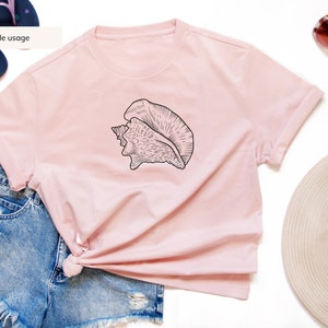 May include: A light pink t-shirt featuring a black seashell graphic. The shirt is styled with denim shorts, sunglasses, a sun hat, and flip-flops. The text "Sample usage" is visible on the image.