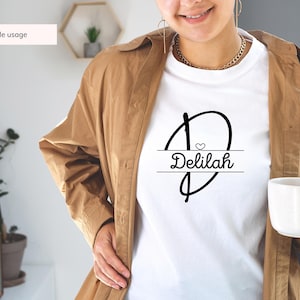 Peut inclure: T-shirt blanc avec un motif graphique noir repr&eacute;sentant une lettre stylis&eacute;e "D" avec un petit c&oelig;ur &agrave; l'int&eacute;rieur et le nom "Delilah" &eacute;crit sous la lettre.