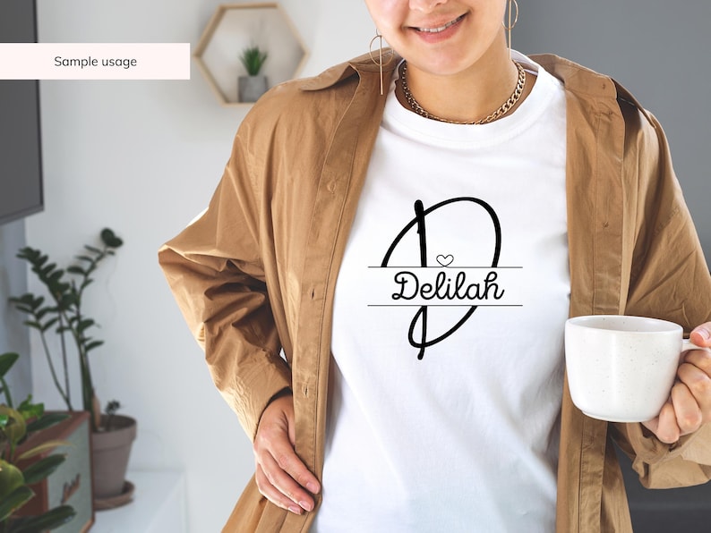 Peut inclure: T-shirt blanc avec un motif graphique noir repr&eacute;sentant une lettre stylis&eacute;e "D" avec un petit c&oelig;ur &agrave; l'int&eacute;rieur et le nom "Delilah" &eacute;crit sous la lettre.