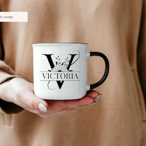 May include: White ceramic mug with a black handle. The mug has a black floral design with the letter "V" and the name "VICTORIA" printed on it.