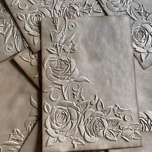 May include: Pale brown paper with an embossed floral rose design. The roses and leaves are arranged in a corner pattern. The paper has a textured appearance, and the embossed details create a three-dimensional effect. Likely used for stationery or crafts.